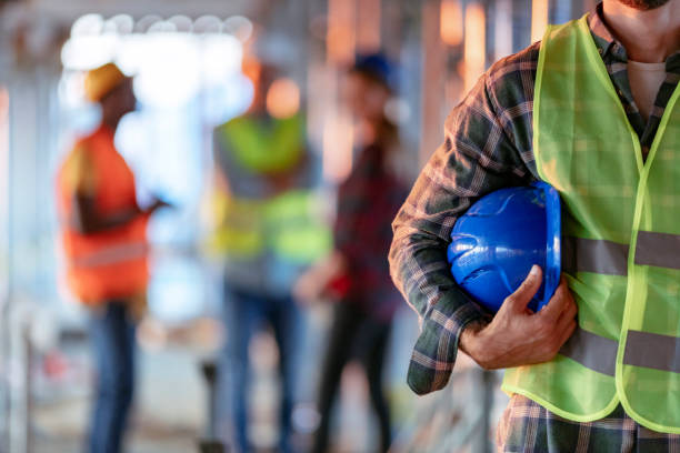 image3 Man holding blue helmet close up. Construction man worker with office and people in background. Close up of a construction worker's hand holding working helmet.